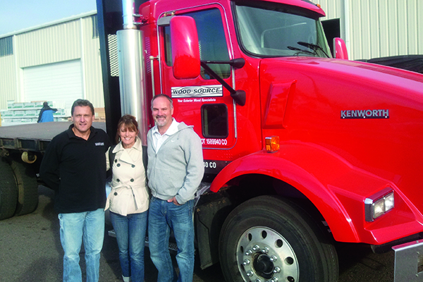 A red semi truck from woodsource, with three people in front.